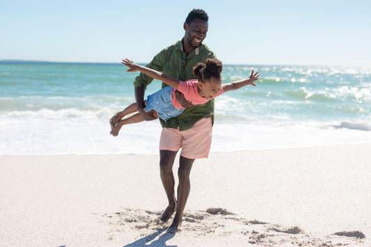 happy african american man carrying daughter pretending to fly at beach on sunny day